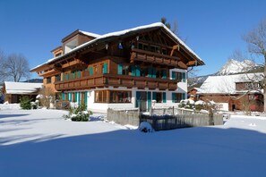 A two-story wooden chalet with a balcony, surrounded by snow.