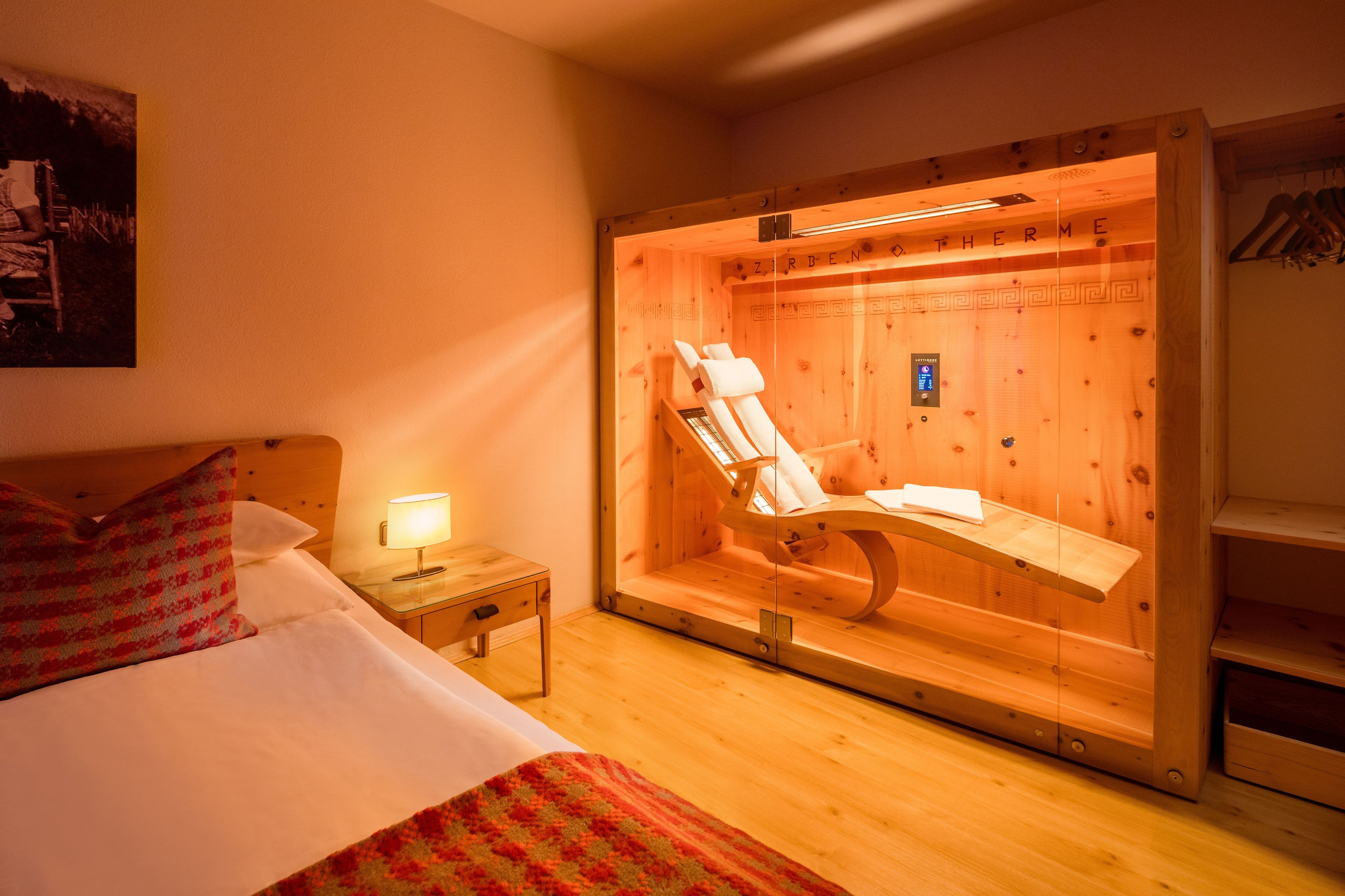 A hotel room with a wooden bed, a small bedside table with a lamp, a wooden sauna, and a wooden shelf with hangers.