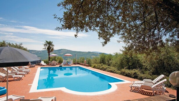 A rectangular swimming pool with a white border, surrounded by lounge chairs and umbrellas, set against a backdrop of hills and trees.