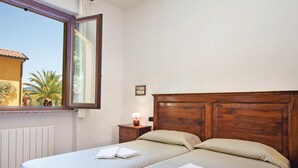 A bedroom with a wooden headboard, two beds, a nightstand with a lamp, a window with a view of palm trees, and a radiator.