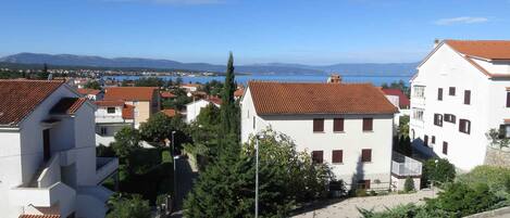 A coastal residential area with white buildings, red-tiled roofs, and a clear view of the sea and mountains in the background.