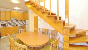 Eine Holztreppe führt zu einer zweiten Etage, eine Küche mit Holzschränken, ein Esstisch mit Stühlen und eine Mikrowelle.