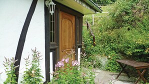 Una cabaña acogedora con puerta de madera, fachada blanca y un jardín con plantas diversas y un banco de madera.