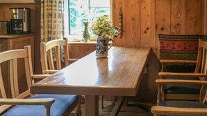 Una mesa de comedor de madera con sillas, una cafetera y un florero con flores en el alféizar de la ventana.