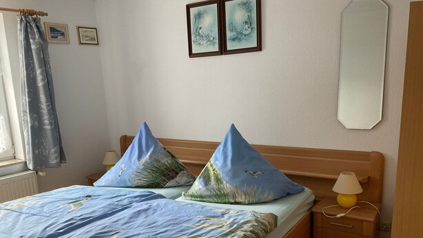 Une chambre à coucher avec un lit, deux oreillers, une table de chevet, une lampe et une fenêtre avec des rideaux.