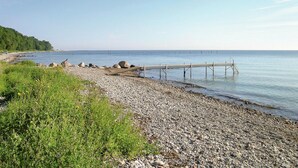 Una playa de guijarros con un muelle de madera que se adentra en aguas tranquilas, rodeada de vegetación y un cielo despejado.