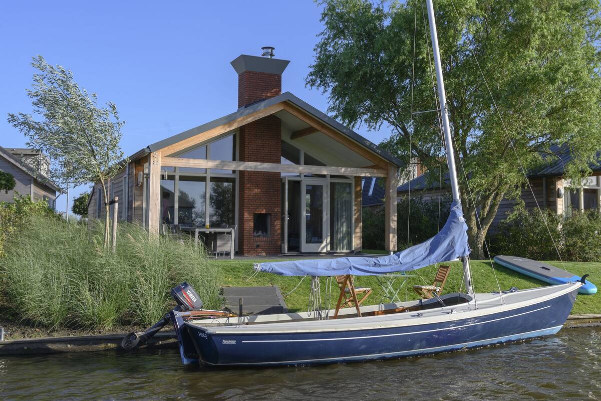 A house with a large glass window, a boat docked in front, and a grassy area.