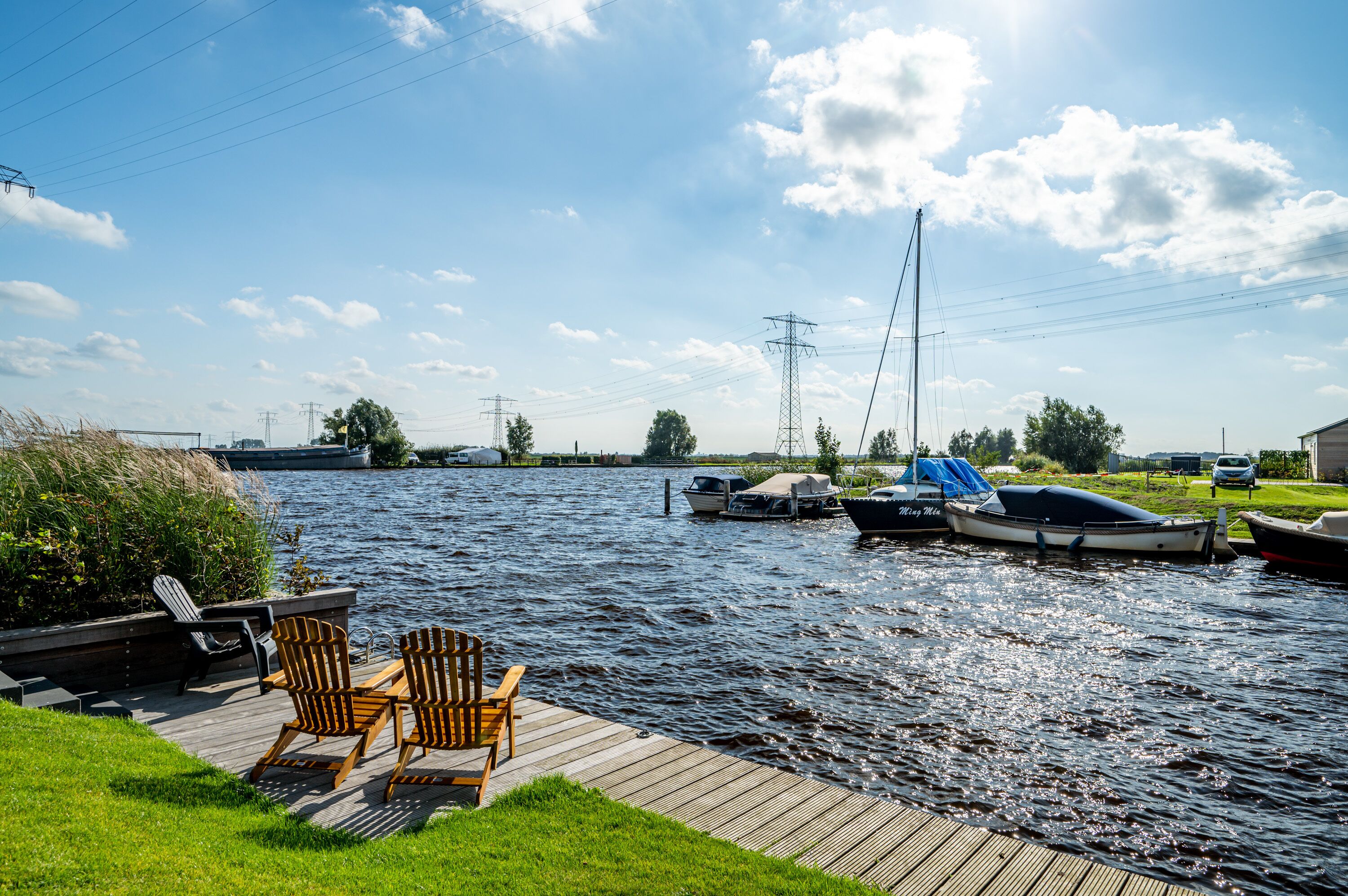 Eine hölzerne Mole mit zwei Adirondack-Stühlen, einem ruhigen Fluss und mehreren Booten, die an der Ufer liegen.
