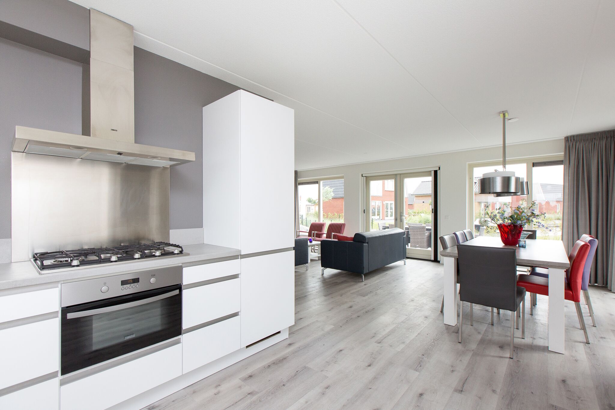 A modern kitchen with a gas stove, oven, and stainless steel range hood. There is a dining area with a table and chairs, and a living room with a sofa and chairs.