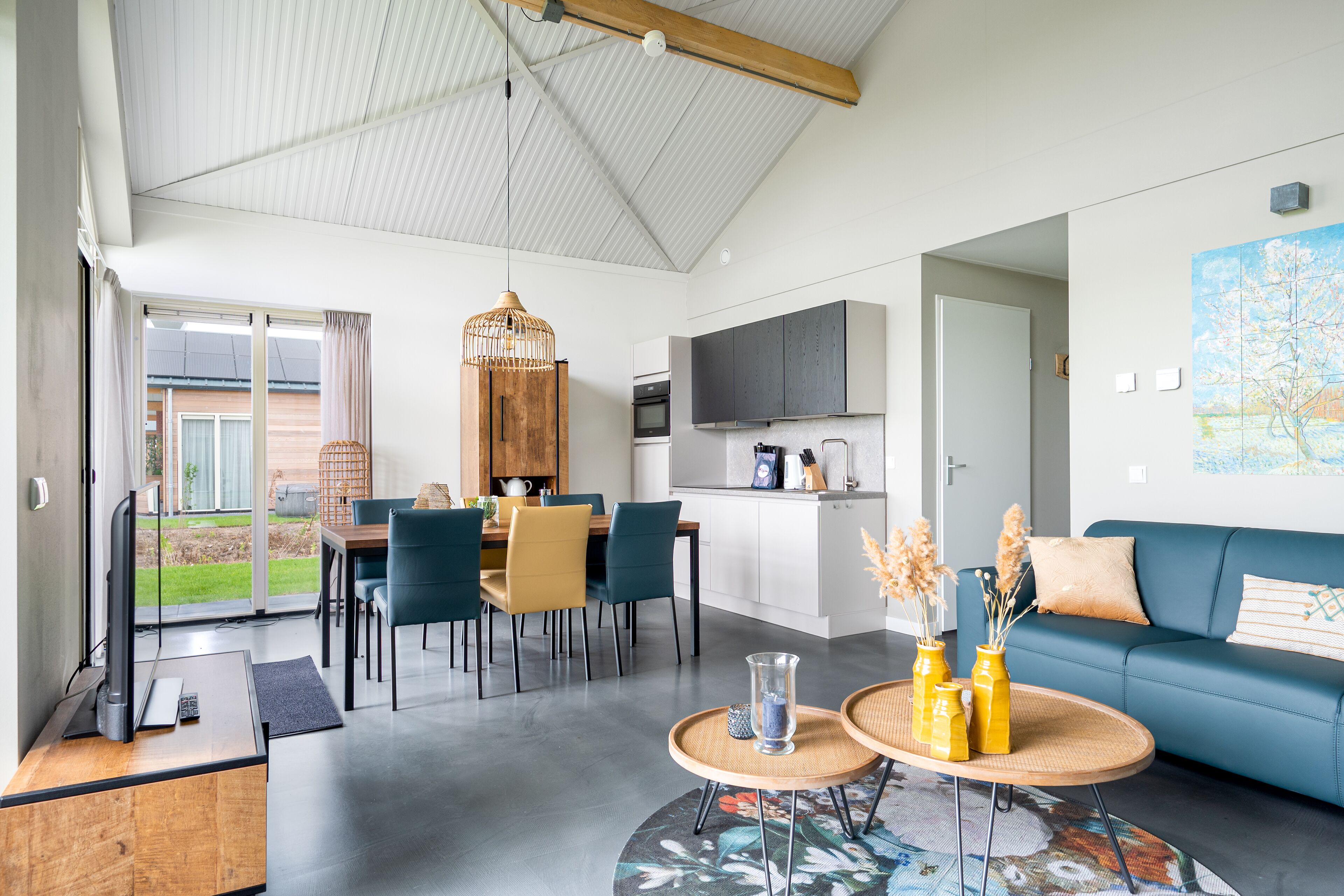 A modern living room with a blue sofa, a dining area with chairs and a table, and a kitchen with cabinets and appliances.