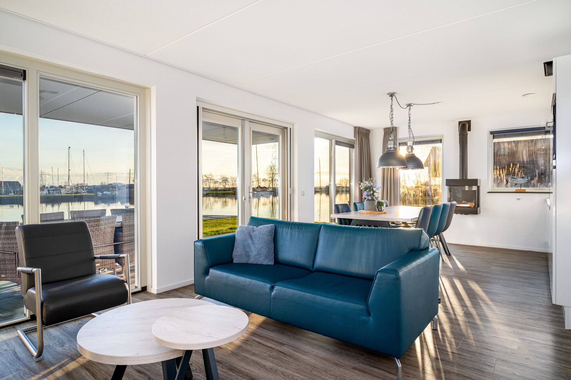 Una sala de estar moderna con un sofá azul, una mesa redonda y vistas a un puerto deportivo a través de amplios ventanales.