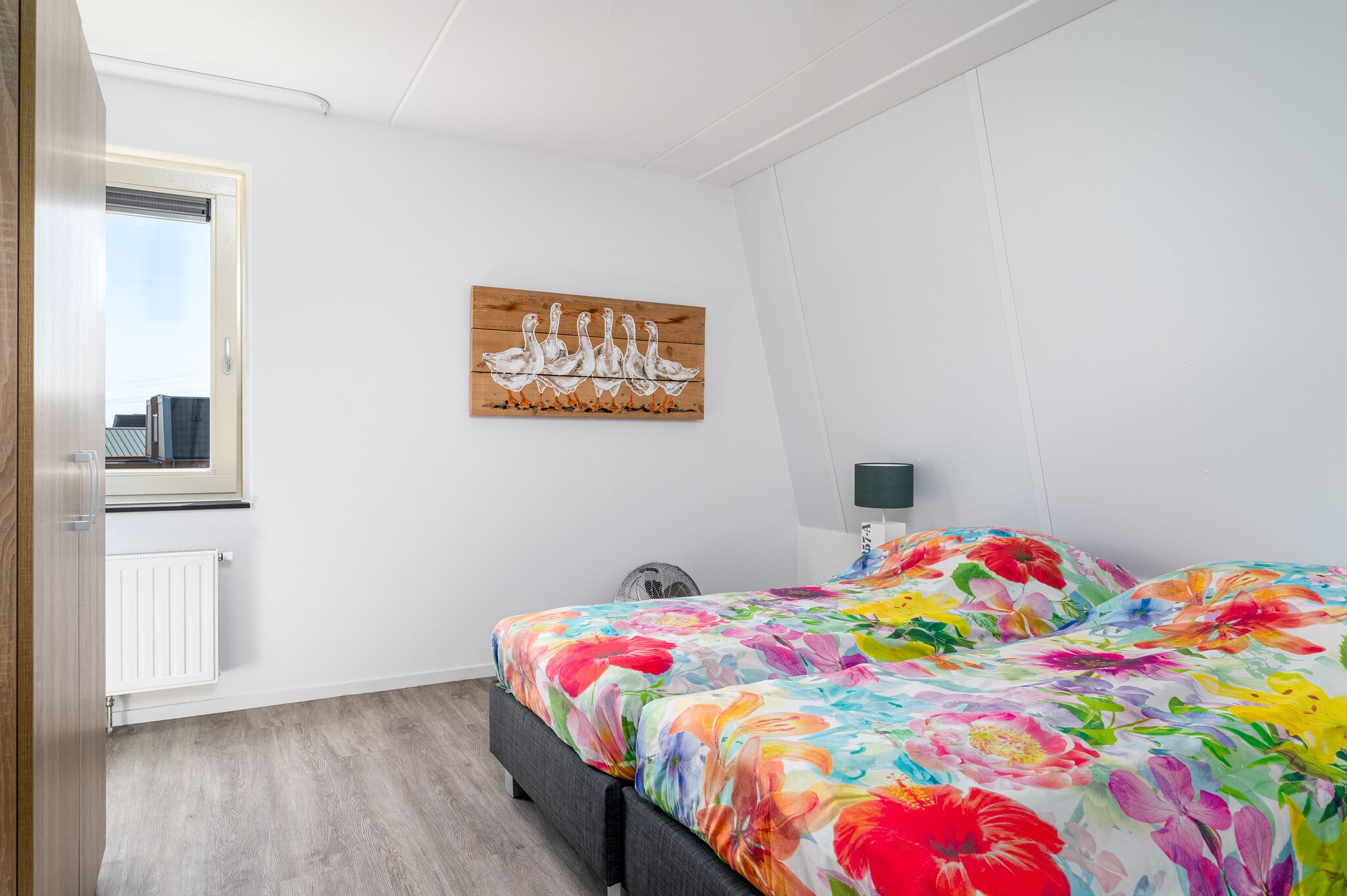 A bedroom with a bed, a window, a radiator, a wall-mounted artwork, and a bedside table with a lamp.