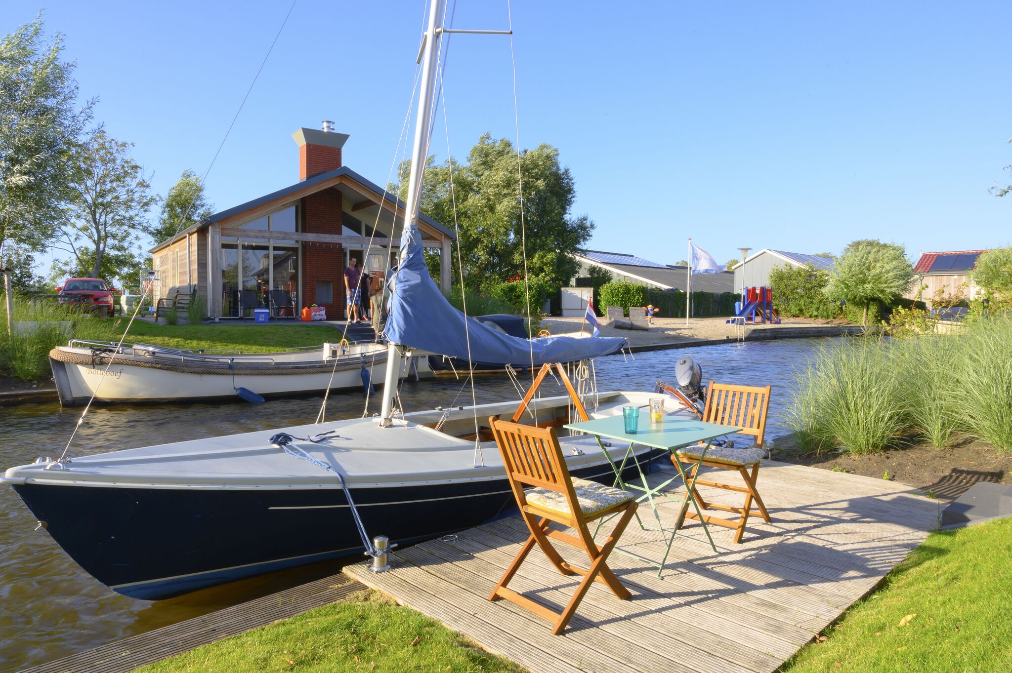 A docked sailboat with a table and chairs on a wooden deck by a canal.