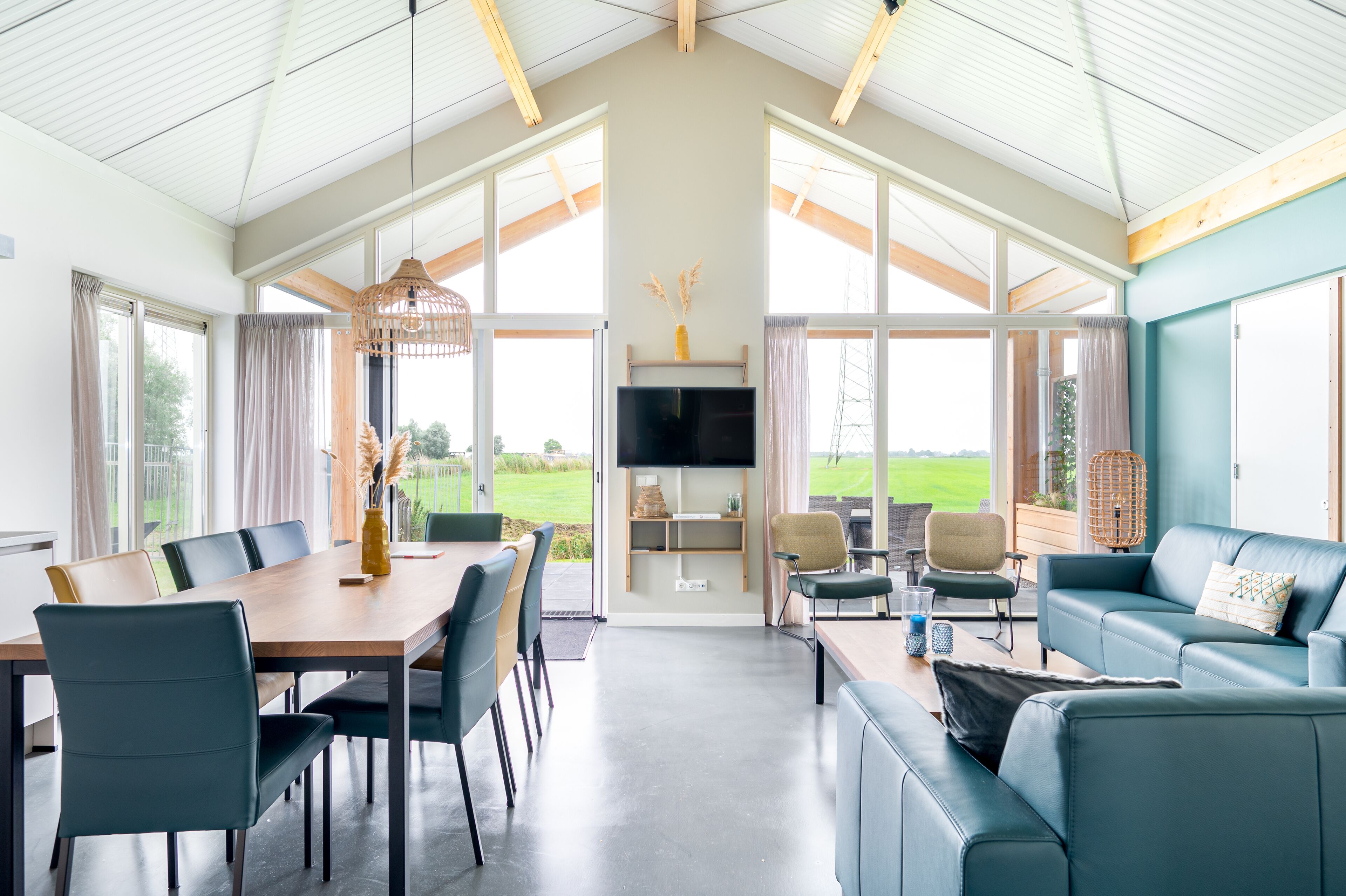 A spacious living area with a dining table, sofas, and a TV. Large windows provide views of the outdoors.