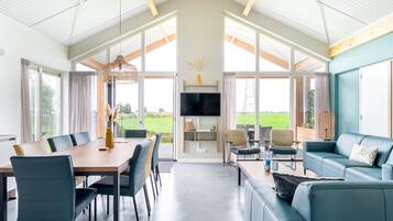 A spacious living area with a dining table, sofas, and a TV. Large windows provide views of the outdoors.