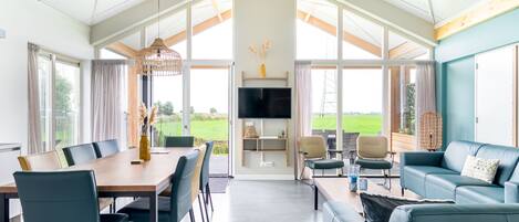 Amplia sala de estar con una mesa de comedor, sofás y televisor. Ventanas grandes ofrecen vistas al exterior.