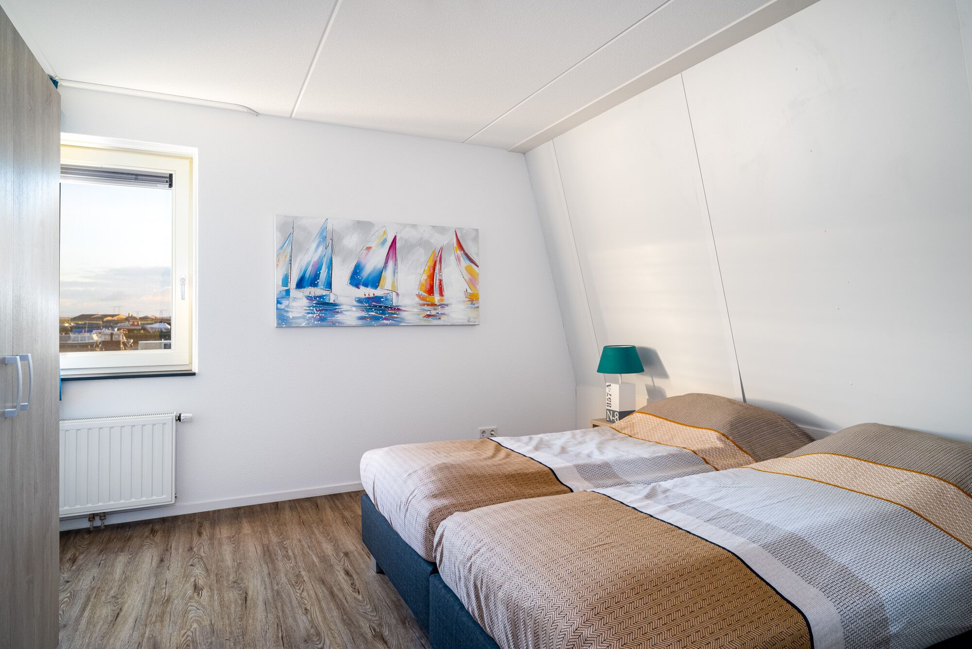Un dormitorio con cama, un cuadro de veleros, una ventana y un radiador.