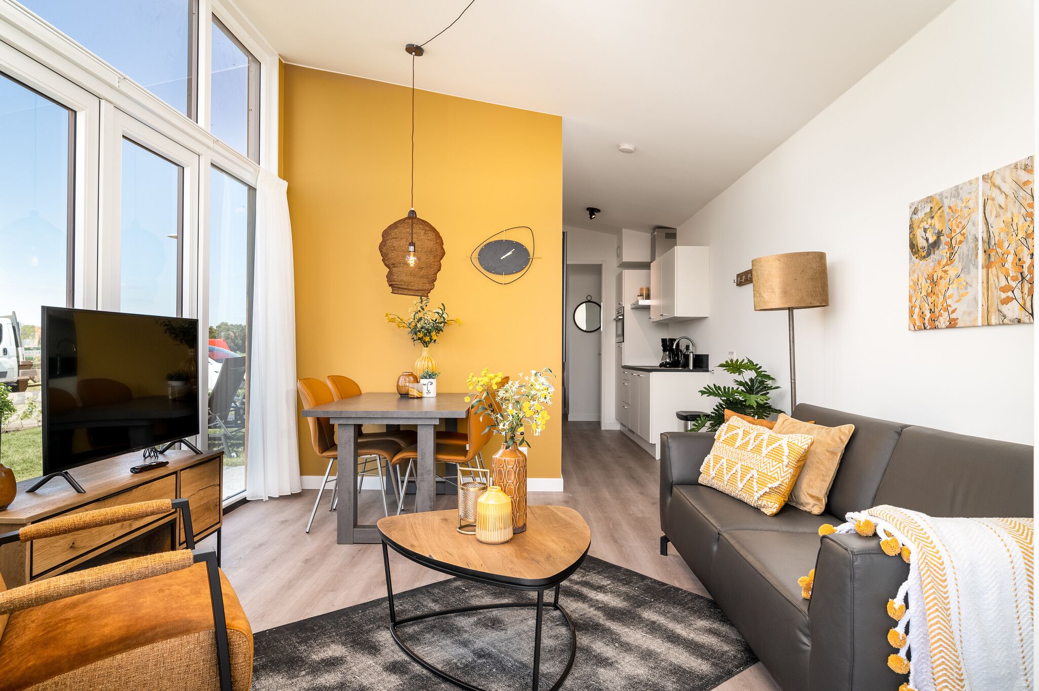 A modern living room with a sofa, coffee table, and TV. There is a dining area with chairs and a table. The room has large windows and a yellow accent wall.