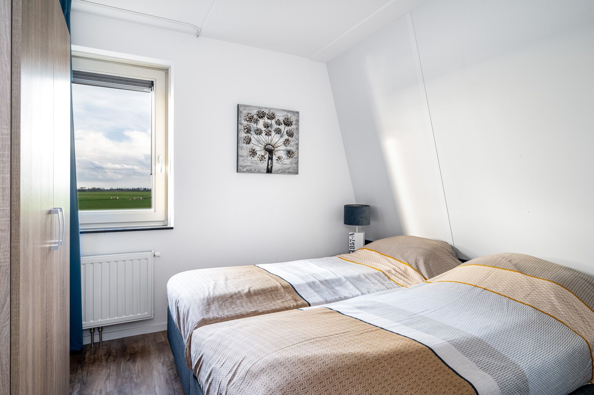 Un dormitorio con cama, un radiador, una ventana con vistas a un campo y una lámpara de pared.
