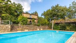 Une piscine aux murs en pierre, une terrasse en bois et un bâtiment en pierre en arrière-plan.