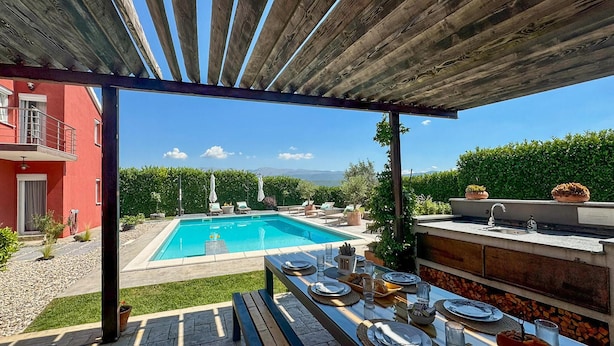 Un espace repas aménagé au bord de la piscine, avec une table dressée pour un repas, un barbecue et une vue sur une piscine et des montagnes en arrière-plan.