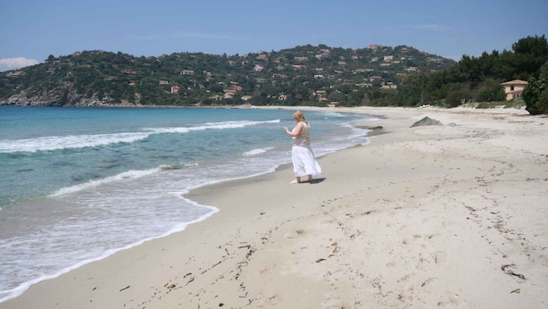 Une plage où l’on voit une personne debout près de l’eau, un rivage sablonneux et une colline avec des bâtiments en arrière-plan.