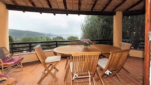 Une terrasse couverte avec une table ronde en bois et des chaises, une plante en pot et une vue sur des montagnes lointaines.