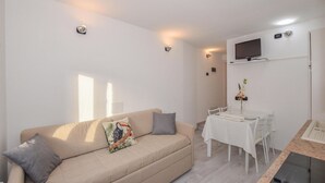 Una sala de estar con un sofá color crema, una mesa de comedor con sillas, un televisor montado en la pared y un pequeño arreglo floral en la pared.