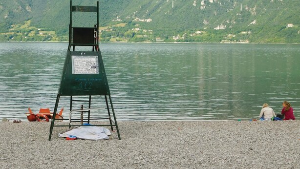 Una torre de salvavidas con un cartel, una playa de guijarros y un lago con montañas al fondo.