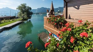 Ein hölzernes Gebäude mit einem Balkon, der auf einen ruhigen Fluss mit viel Grün und Blumen blickt.