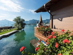 Ein hölzernes Gebäude mit einem Balkon, der auf einen ruhigen Fluss mit viel Grün und Blumen blickt.