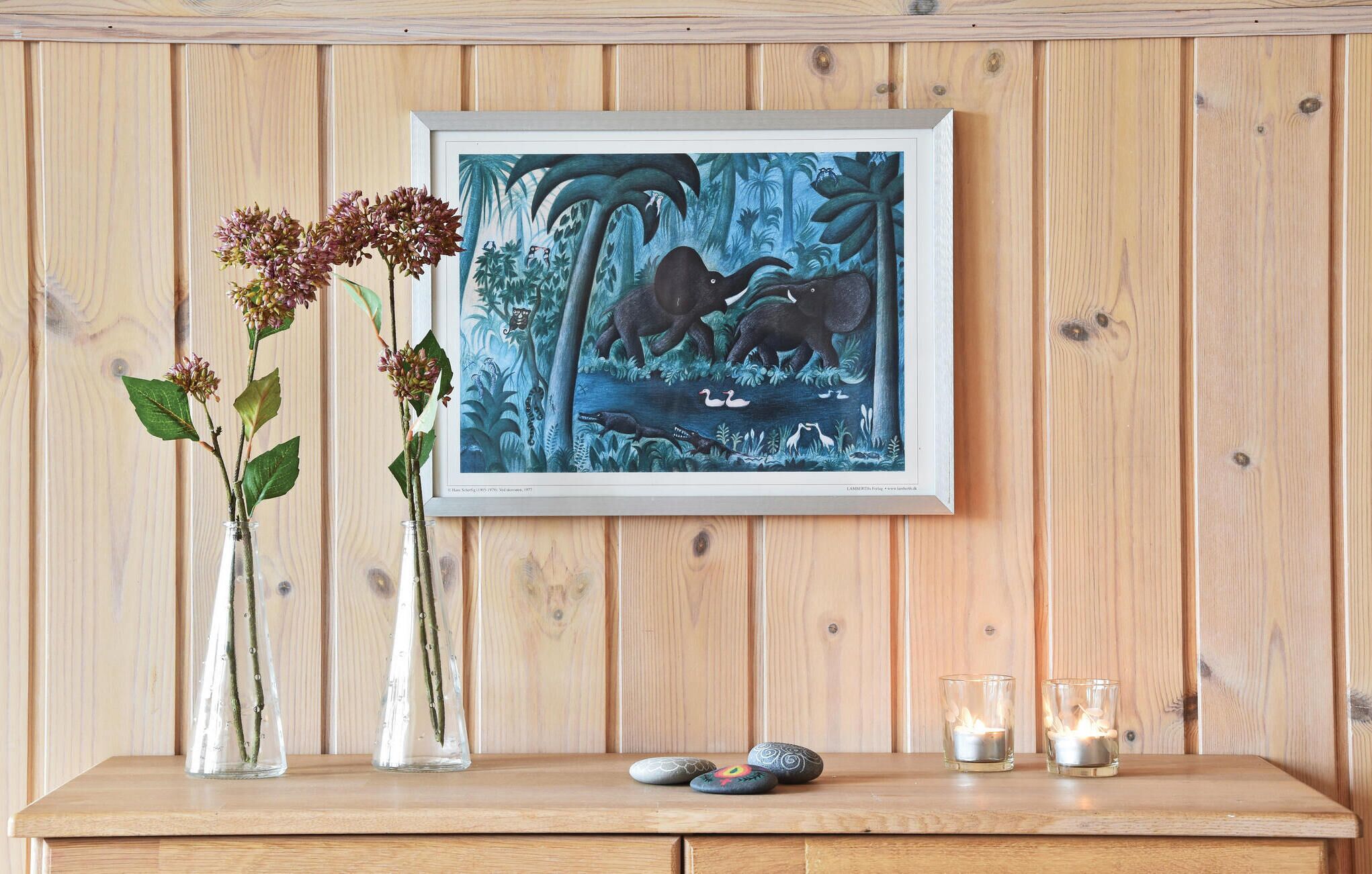 Una obra de arte enmarco con elefantes y pájaros sobre una pared de madera, un jarrón con flores y dos velas encendidas sobre una mesa de madera.
