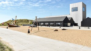 A modern building with a large glass facade, a concrete structure, and a sandy beach area with a wooden ship model. - Lovely home in Ulfborg with WiFi (Ulfborg)