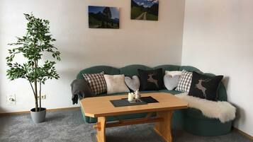 A cozy living room with a green sofa, a wooden coffee table, a potted plant, and two wall-mounted pictures.