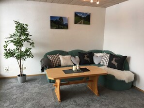 A cozy living room with a green sofa, a wooden coffee table, a potted plant, and two wall-mounted pictures.