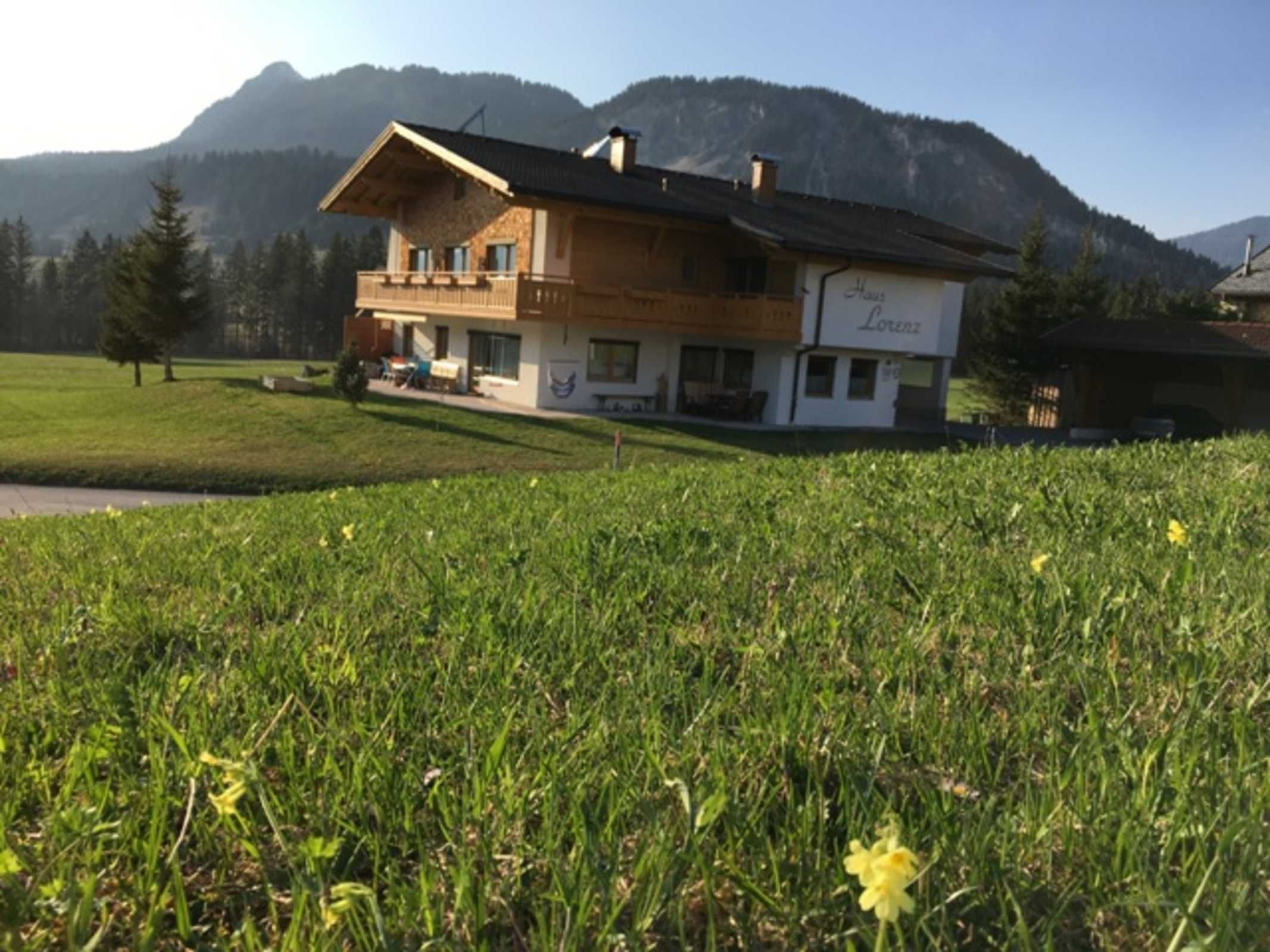 A two-story house with a balcony, surrounded by a grassy field and mountains in the background.