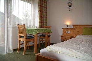A room with a bed, a wooden chair, a table with a green tablecloth, a nightstand, and a wall-mounted lamp.