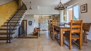 Une cuisine spacieuse avec un mur en pierre, une table à manger en bois et des chaises. Un escalier avec une rampe en métal mène à un autre niveau.
