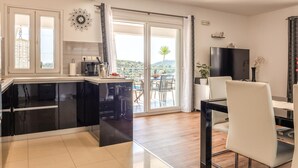 Une cuisine moderne dotée d’un grand îlot central, d’un coin repas avec une table et des chaises, et d’une porte-fenêtre coulissante donnant sur un balcon avec vue sur les collines.