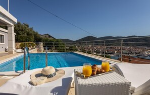 Une piscine avec vue sur la ville et les montagnes, un fauteuil de relaxation, un chapeau de soleil et un plateau avec des croissants et du jus d’orange.