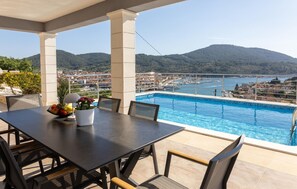 Une terrasse avec une table et des chaises, une piscine et une vue sur un port de plaisance et des collines.