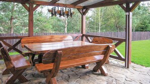 Een houten picknicktafel met banken onder een houten pergola, op een terras van stenen, met een houten omheining en bomen op de achtergrond.