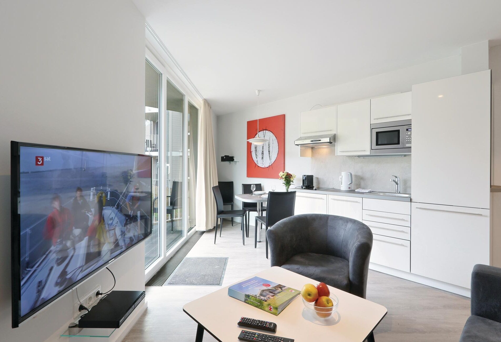 Ein modernes Wohnzimmer mit Flachbildfernseher, einem Essbereich mit Tisch und Stühlen, einer Küche mit weißen Schränken und Edelstahlgeräten sowie einem großen Fenster mit Vorhängen.