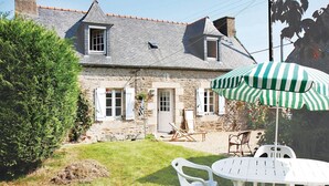 A stone house with a sloped roof, a small garden with a white plastic table and chairs, and a green and white striped umbrella. - Awesome home in Pleumeur Gautier (Pleumeur Gautier)
