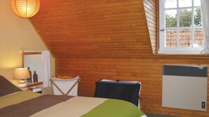 A bedroom with a sloped ceiling, a bed with a striped bedspread, a bedside lamp, a small table with a bottle and books, a window with a view of a brick wall, and a wall-mounted heater. - Awesome home in Pleumeur Gautier (Pleumeur Gautier)