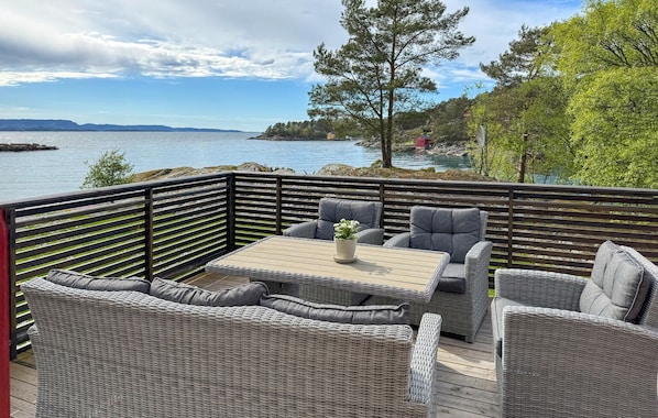 A deck with wicker furniture, a table, and a view of a lake and distant mountains.