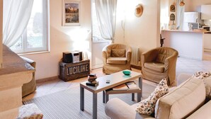 A cozy living room with two beige armchairs, a wooden coffee table, and a sofa. A vintage radio is on a wooden crate labeled 'LUCILINE'. A framed picture hangs on the wall, and a window with white curtains is visible. - 2 bedroom lovely home in Trie-Château (Trie-Château)