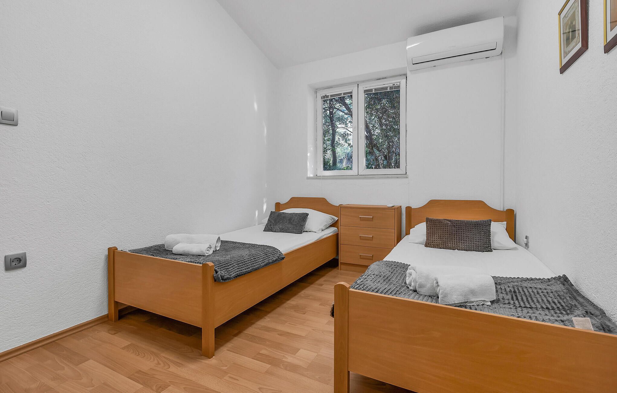 A small, clean room with two wooden beds, a small dresser, and a window with a view of trees.