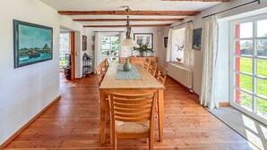 Ein Esszimmer mit Holztisch und -stühlen, einer Hängeleuchte und großen Fenstern mit Vorhängen.