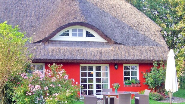 Ein Haus mit einem Reetdach, rotem Anstrich, einem weißen Fenster und einer Terrasse mit Gartenmöbeln.